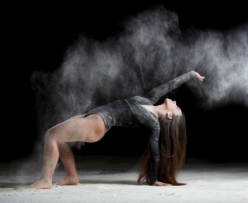 young beautiful caucasian woman with long hair is dressed in a sports black bodysuit stands in a bent pose and throws white flour, black background, gymnastic exercises
