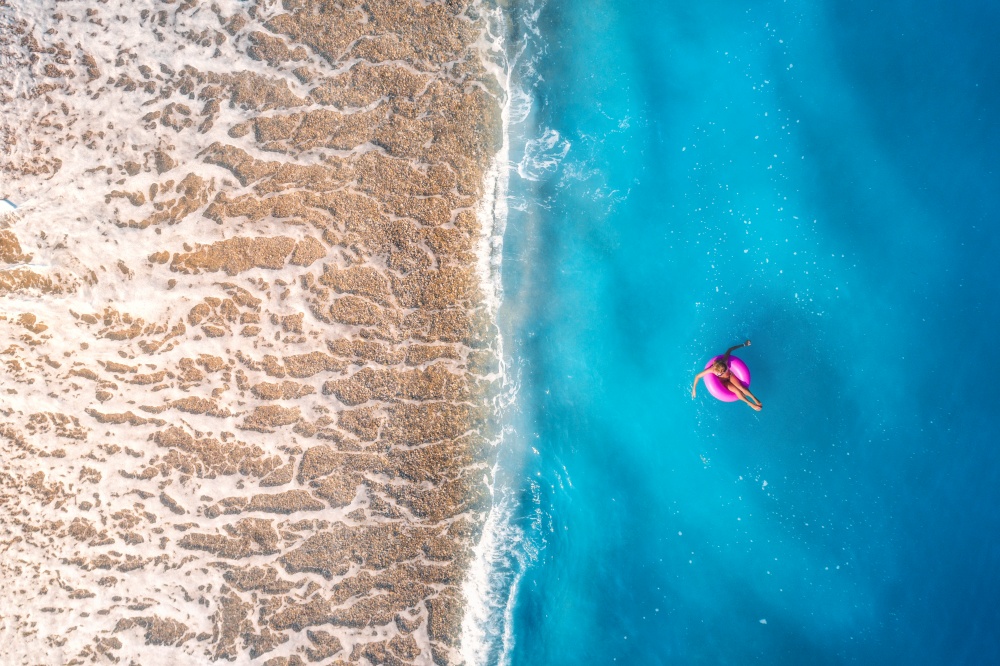 Aerial view of a young woman swimming with pink donut swim ring in clear blue sea with waves at sunset in summer. Tropical landscape with girl, azure water, sandy beach. Top view. Travel and tourism. Aerial view of a young woman swimming with pink donut swim ring