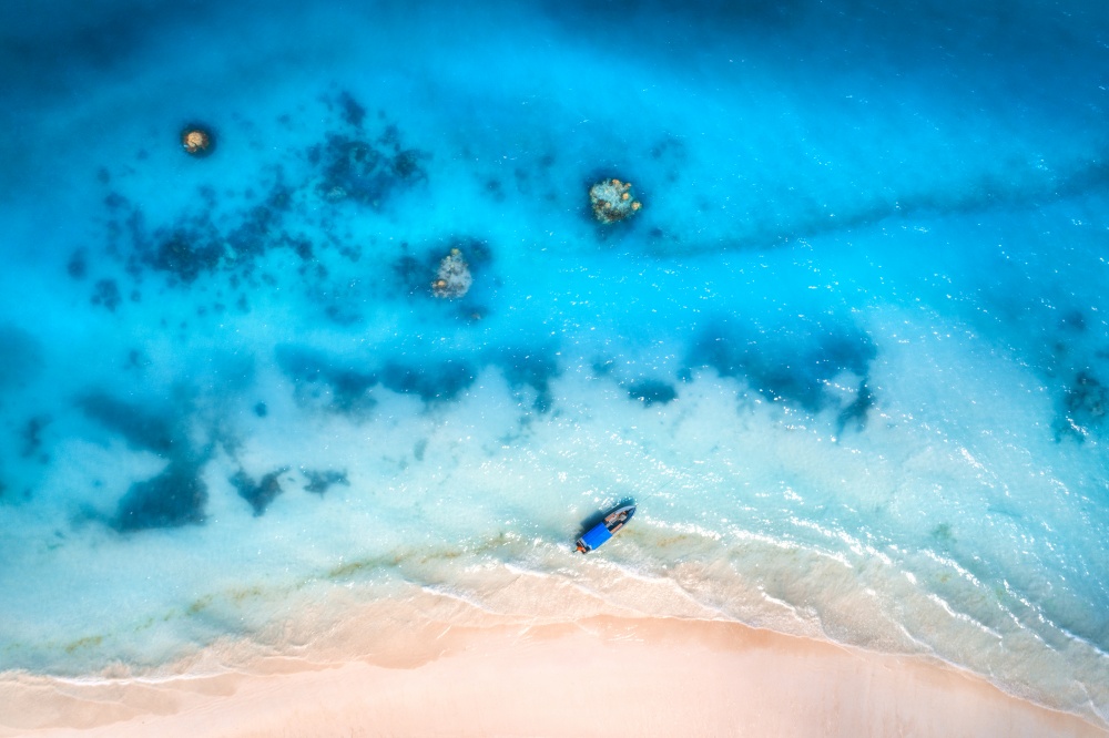 Aerial view of alone fishing boat in blue transparent water at sunset in summer. Top view of boat, yacht, white sandy beach in Zanzibar. Travel. Tropical seascape with sailboat, sea. View from above. Aerial view of the fishing boat in clear blue water at sunset