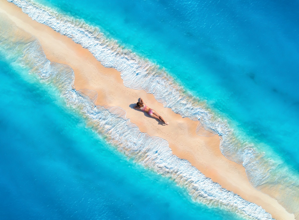 Aerial view of the beautiful young lying woman on the sandy beach and blue sea with waves on the both sides at sunset. Summer holiday. Top view of the back of sporty slim girl, sandbank, clear water. Aerial view of the beautiful young lying woman on the sandy beac