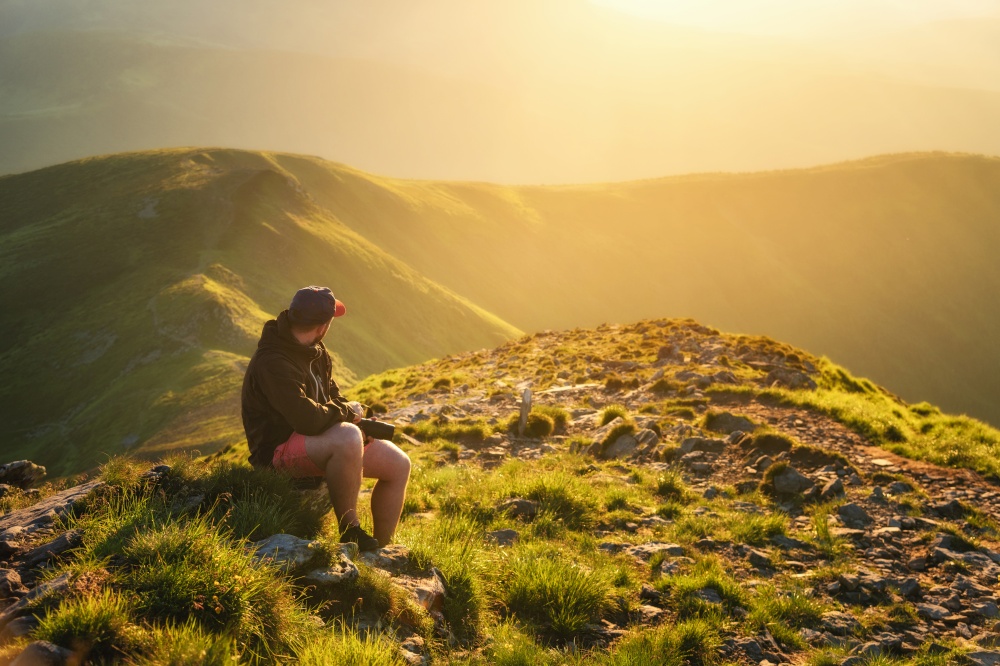 Beautiful mountains in fog and young man is sitting on the stone on mountain peak at sunset in summer. Landscape with relaxing guy on the trail, green grass, hills lighted by sunbeams. Travel