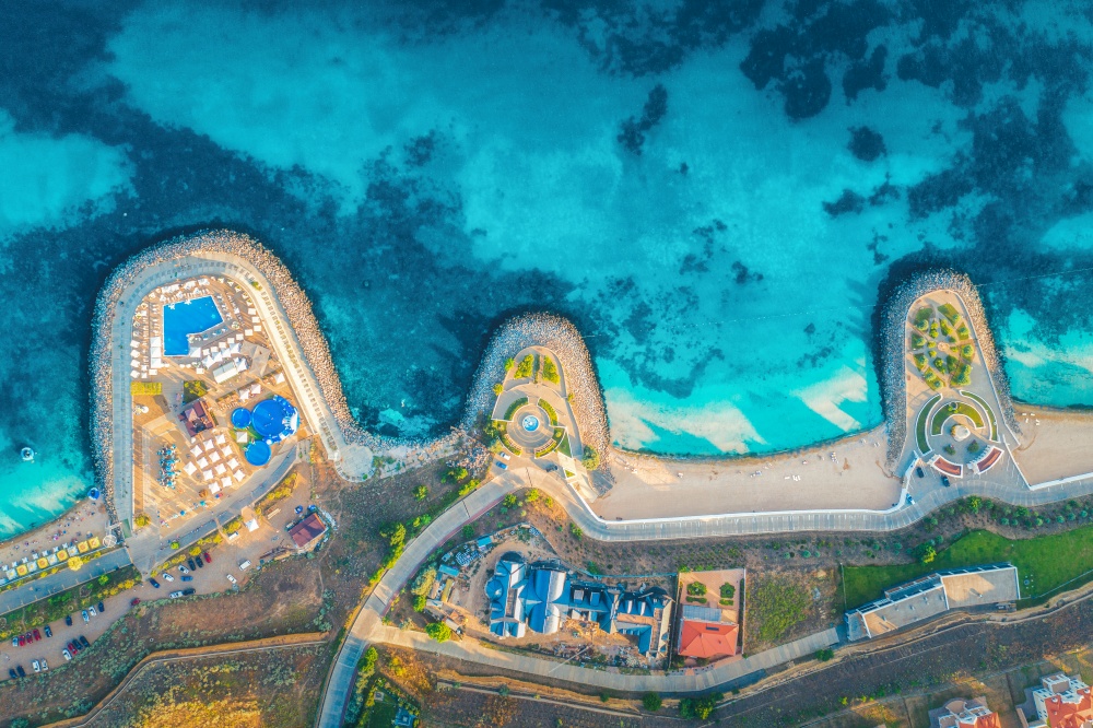 Aerial view of beautiful sandy beach, blue sea, restaurants on the promenade, pool, umbrellas, swimming people in clear water, green trees at sunset in summer. Top view of seafront. Tropical landscape