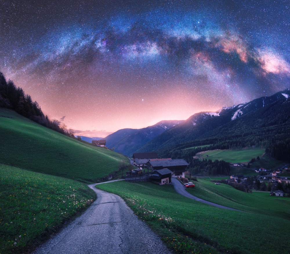 Arched Milky Way over the rural mountain road in summer in Italy. Beautiful night landscape with starry sky, milky way arch, winding road in mountain village, hills, green meadows and buildings. Space. Arched Milky Way over the rural mountain road in summer in Italy