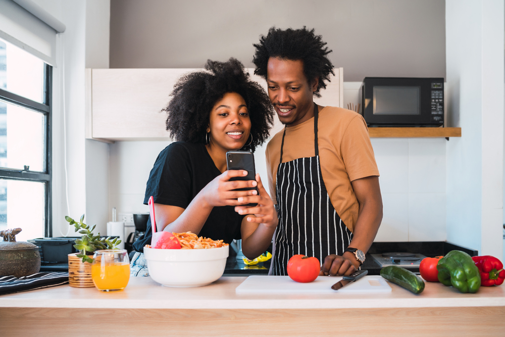Portrait of young afro couple cooking together and using mobile phone in the kitchen at home. Relationship, cook and lifestyle concept.