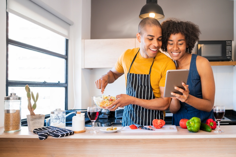 Portrait of young latin couple using a digital tablet and smiling while cooking in kitchen at home. Relationship, cook and lifestyle concept.