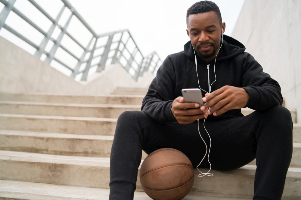 Portrait of an athletic man using his mobile phone on a break from training while sitting on concrete stairs. Sport and health concept.