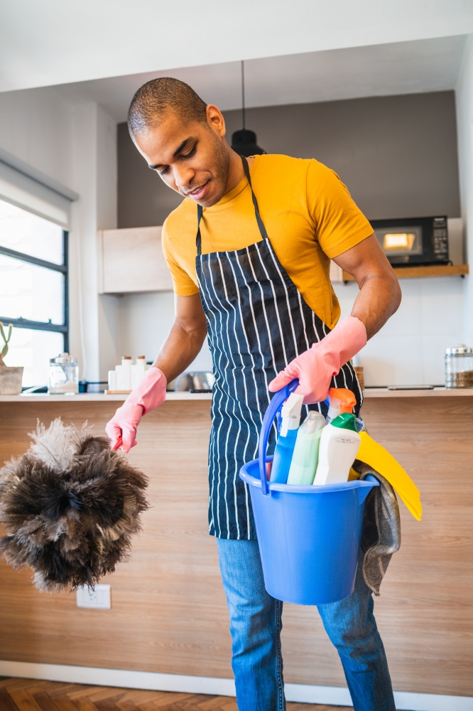 Portrait of young latin man holding a bucket with cleaning items at home. Housekeeping and cleaning concept.