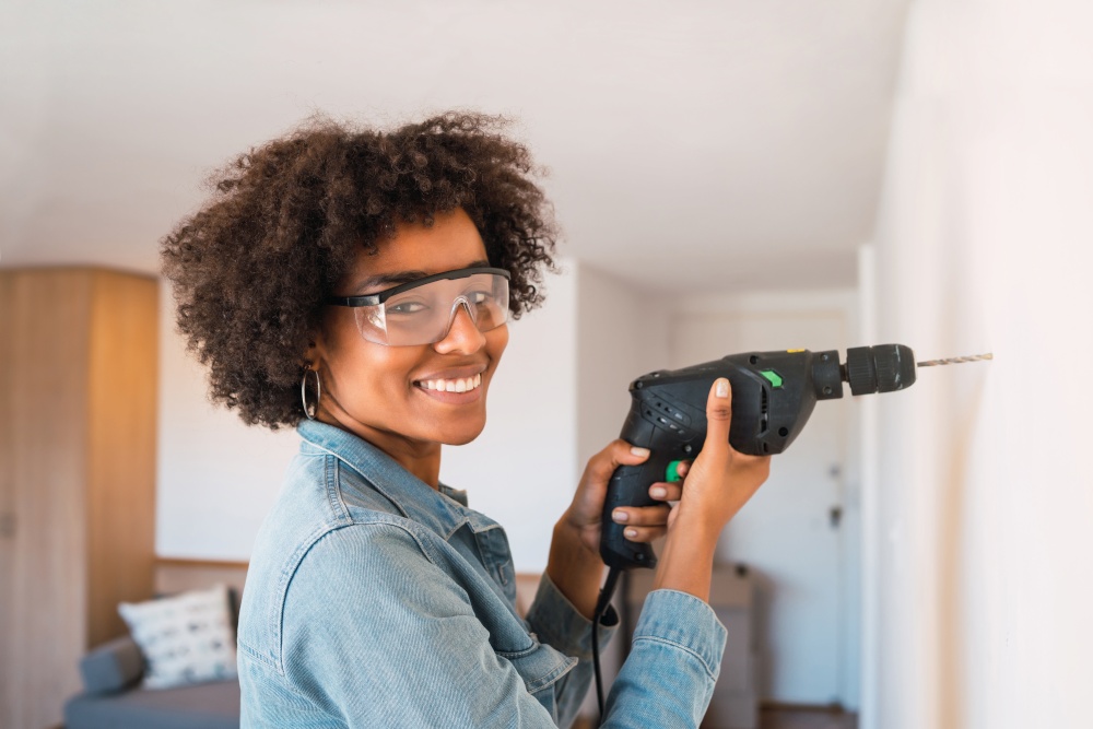 Portrait of young afro woman drilling wall with an electric drill at home. Home improvement concept.
