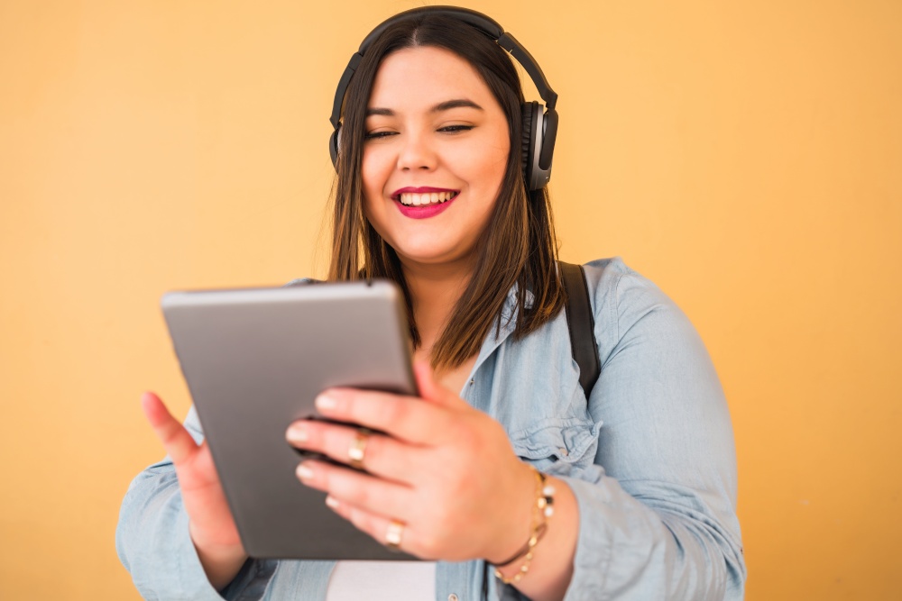 Portrait of young plus size woman listening to music with headphones and digital tablet outdoors against yellow background.