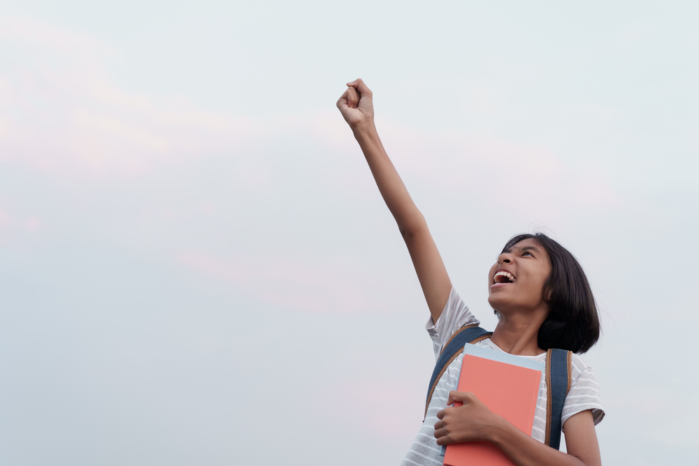 Happy Asian schoolgirl success and excited on the face while holding a book with hand raised up to blue sky background  in the summer at morning