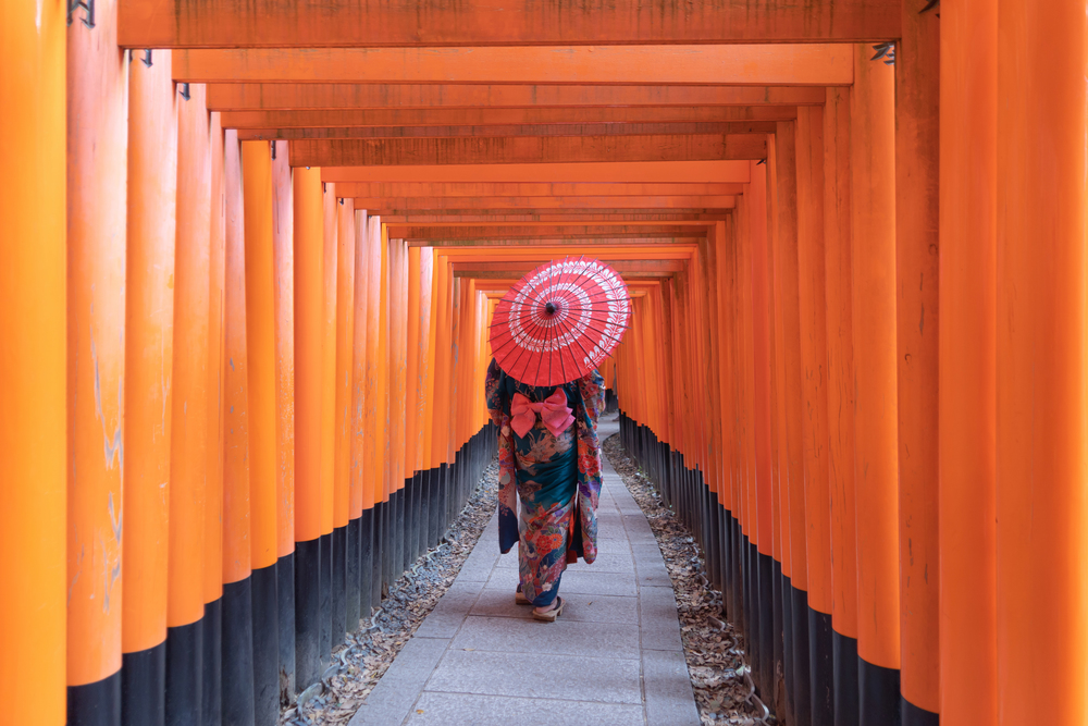 An Asian woman wearing Japanese traditional kimono standing in Fushimi Inari Taisha during travel holidays vacation trip outdoors in Kyoto, Japan. Red poles in the temple. Walkway tunnel of shrine.