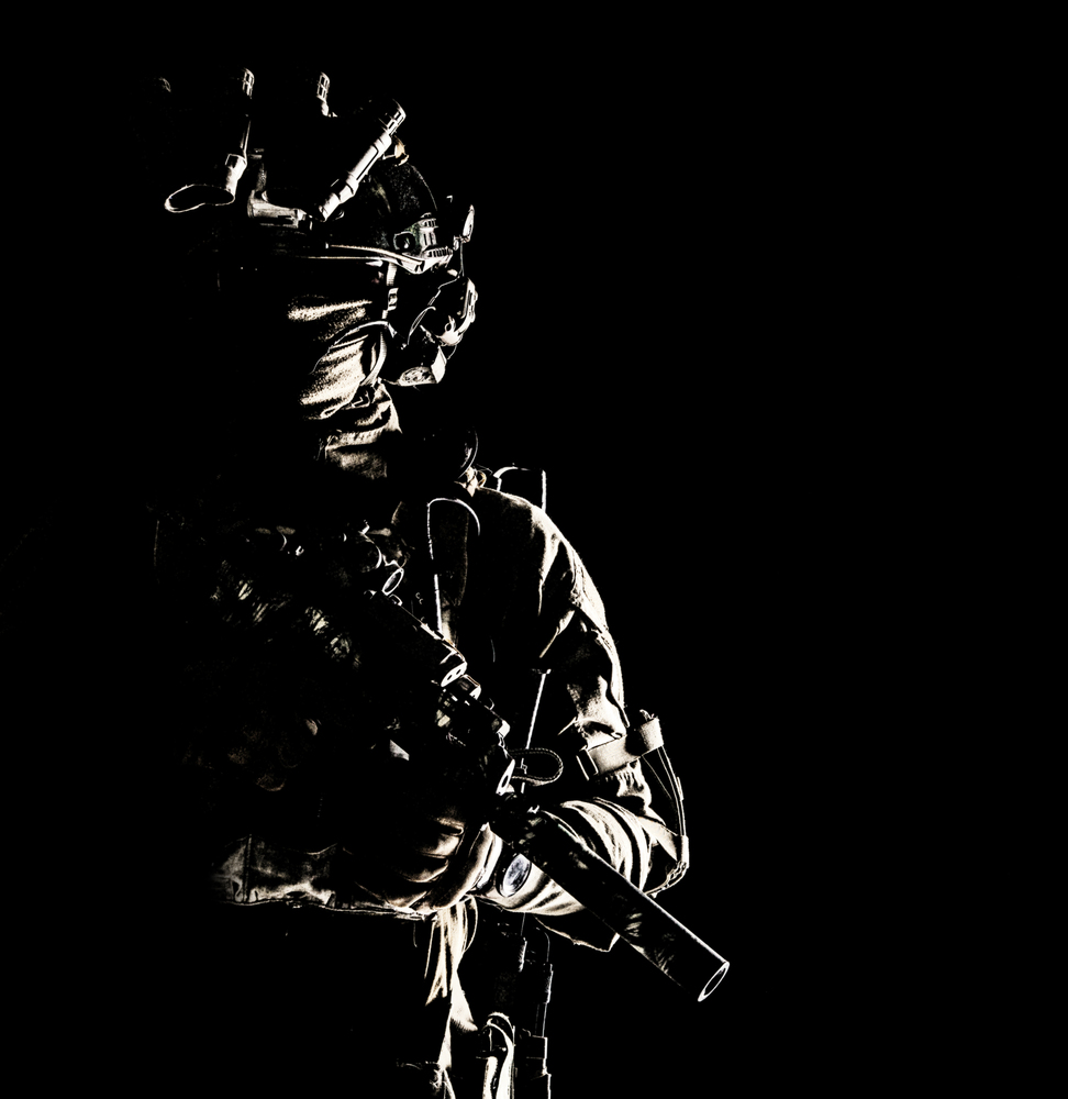 Army special operations forces soldier in mask and combat uniform, helmet equipped night-vision device, armed submachine gun with silencer, looking aside, low key studio portrait on black, copyspace. Special operations forces soldier low key portrait