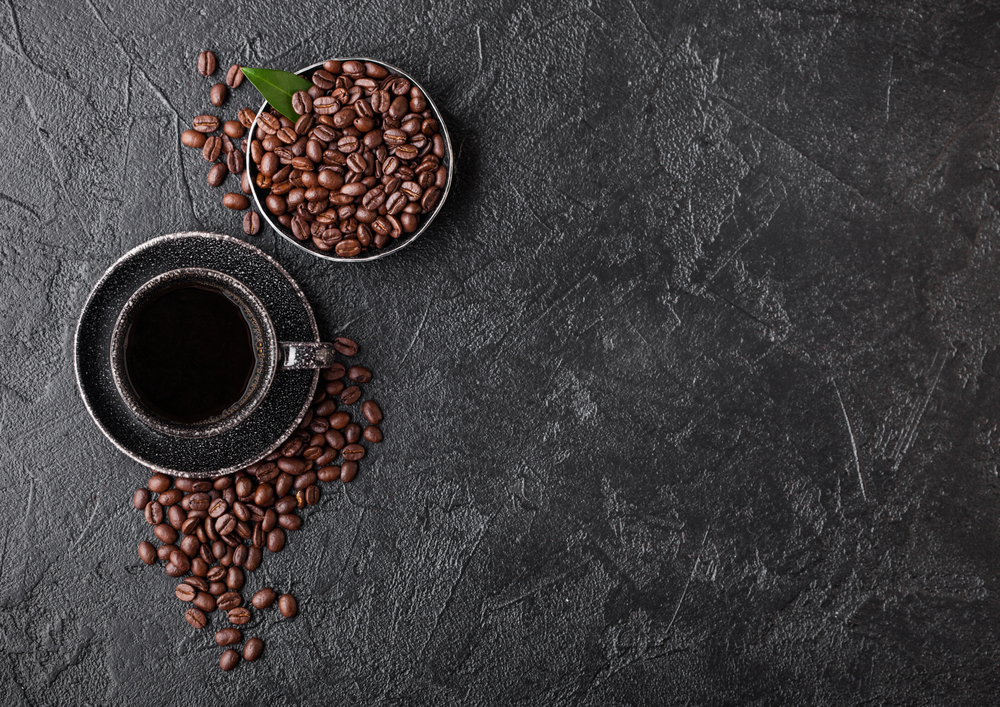 Cup of fresh raw organic coffee with beans and coffee tree leaf on black background. Black ceramic mug.