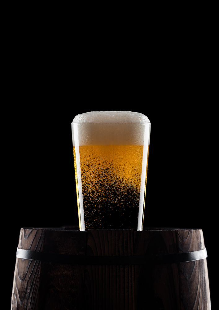 Cold glass of craft beer on old wooden barrel on black background with dew and bubbles