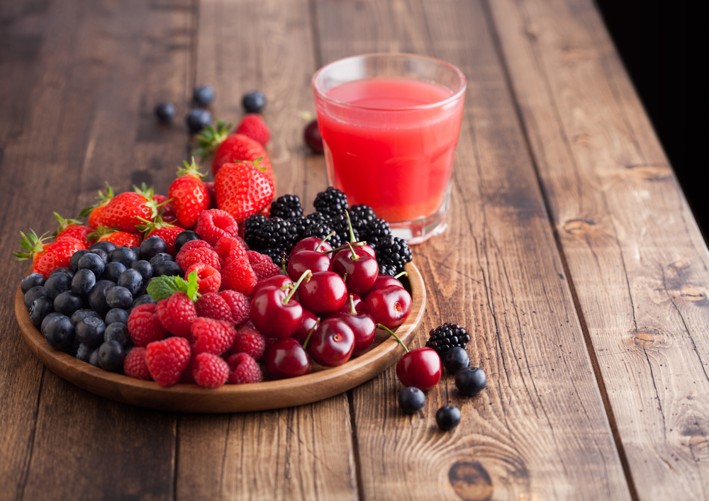 Fresh organic summer berries mix in round wooden tray with glass of juice on light wooden table background. Raspberries, strawberries, blueberries, blackberries and cherries.  Top view