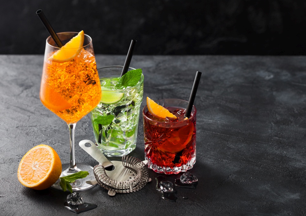 Glasses of spritz,mojito and negroni cocktails with ice cubes and lime and orange slices with mint leaf and black straw on dark background with strainer. Top view