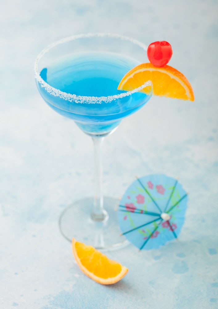 Blue lagoon summer cocktail in margarita glass with sweet cocktail cherries and orange slice with umbrella on blue table background. Top view