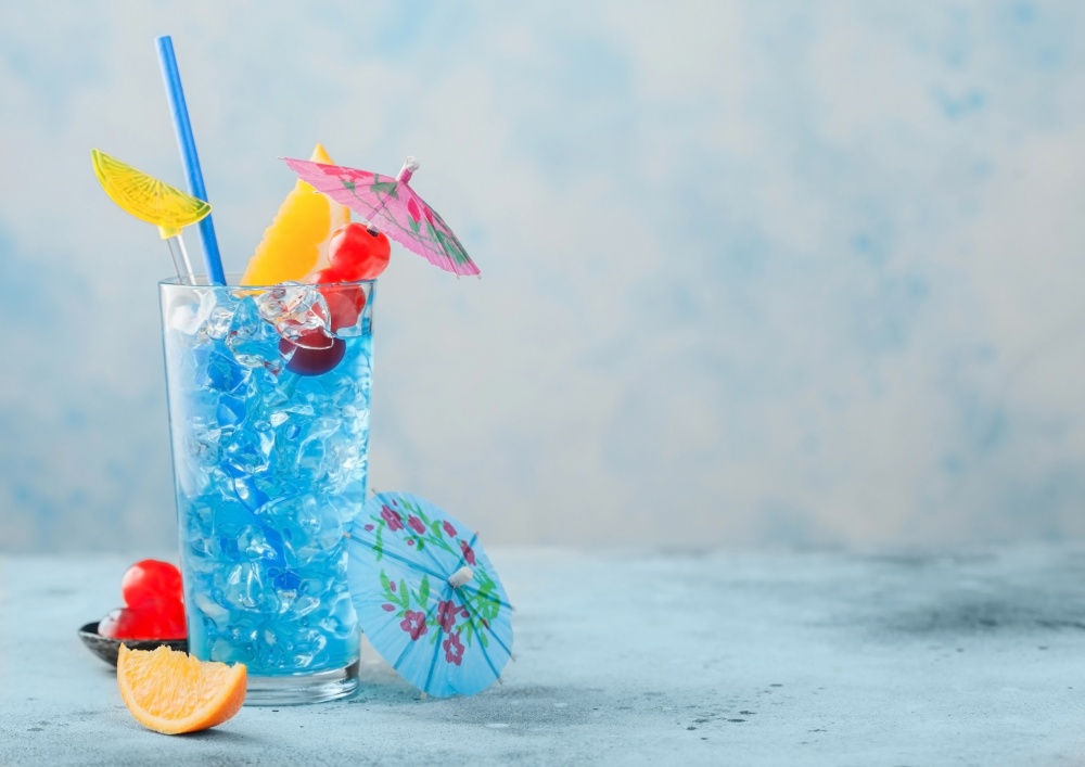 Blue lagoon summer cocktail in highball glass with sweet cocktail cherries and orange slice with umbrella on blue table background. Space for text