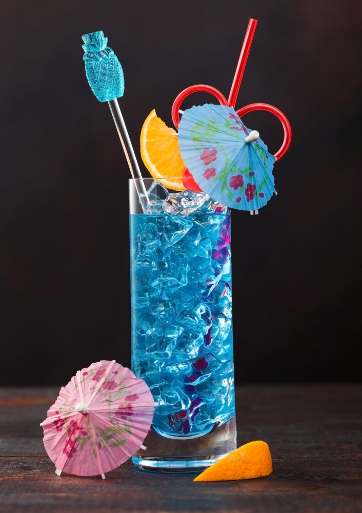 Blue lagoon summer cocktail in highball glass with sweet cocktail cherries and orange slice with umbrella on dark table background.