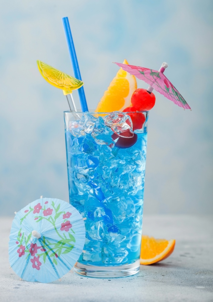 Blue lagoon summer cocktail in highball glass with sweet cocktail cherries and orange slice with umbrella on blue table background.