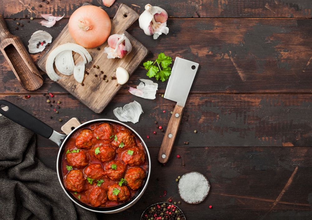 Beef traditional meatballs in tomato sauce in frying pan with pepper, garlic and parsley with onion and cleaver on wooden board. Top view