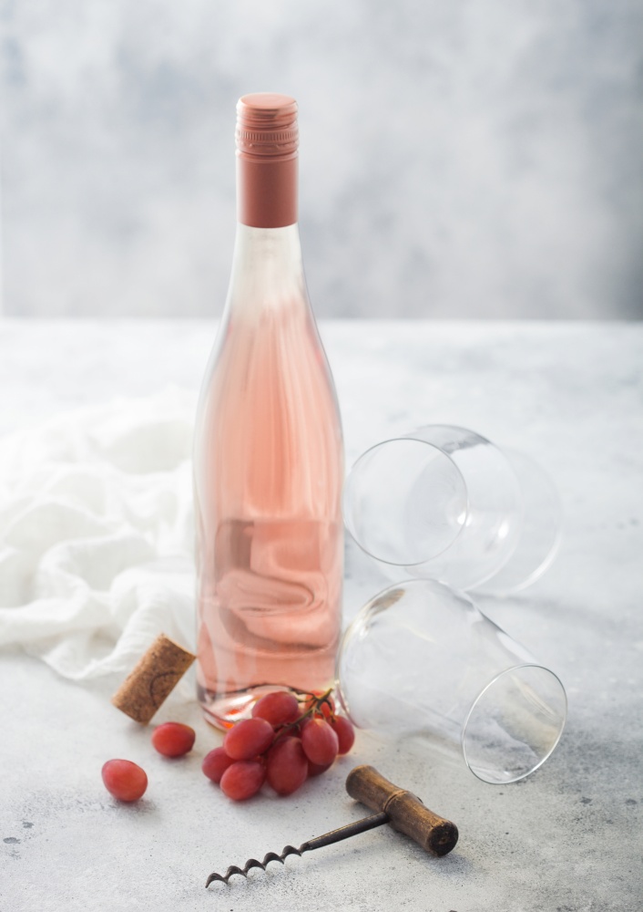 Bottle of pink rose wine with grapes and empty glass with corkscrew and linen cloth on light table background.