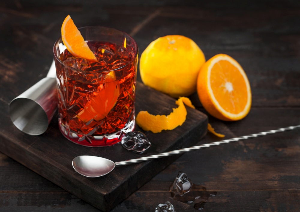 Negroni cocktail in crystal glass with orange slice and fresh raw oranges on chopping board with spoon on wooden background. Ice cubes and jigger.
