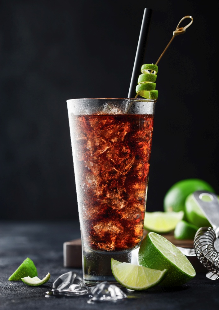 Cuba Libre cocktail in highball glass with ice and lime peel on bamboo stick with straw and fresh limes on black table background.