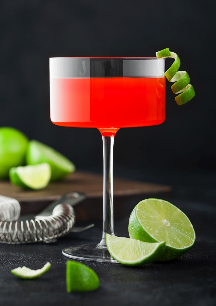 Cosmopolitan cocktail in modern crystal glass with lime peel and fresh limes with strainer on black table background.
