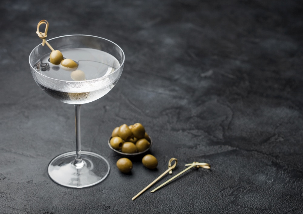 Vodka martini gin cocktail in modern glass with olives in metal bowl and bamboo sticks on black background.Top view