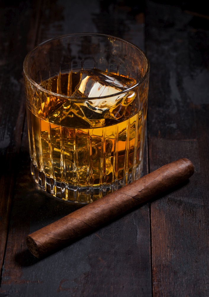 Single malt scotch whiskey in crystal glasses with ice cubes and cuban cigar on wooden table background.