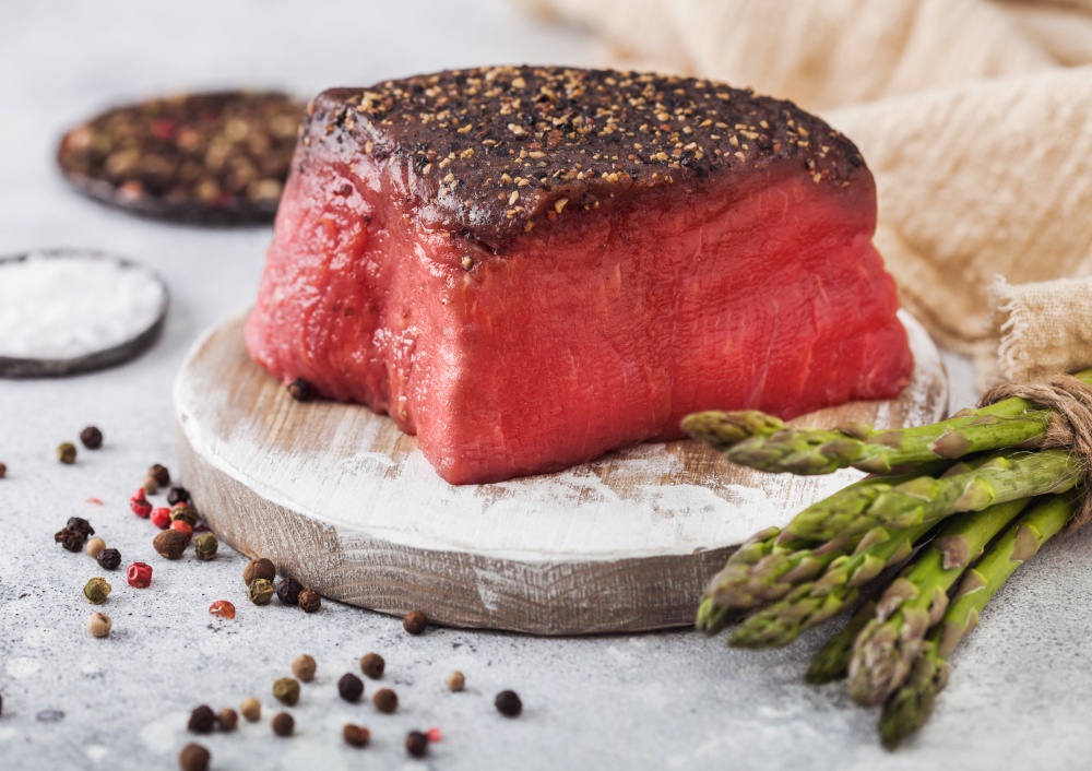 Slice of Raw Beef Topside Joint with Salt and Pepper on round chopping board with asparagus tips and garlic on light kitchen table background.