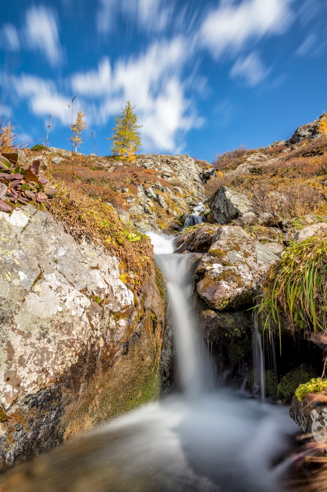 View of a waterfall with smooth water, blue sky and flowing clouds in the background in Altai mountains, Siberia, Russia. Fall 2019