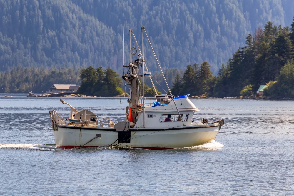 Shot of a commercial fishing boat sailing in a harbour in Sitka, AK. Green forest and a house in the background.