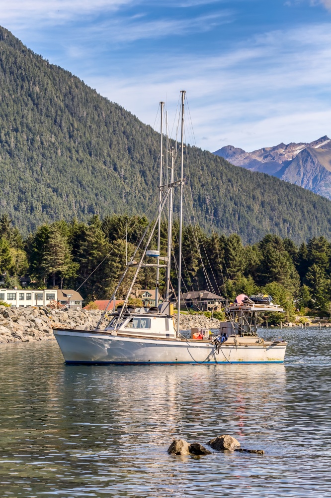 Shot of a fishing boat sailing in a harbour in Sitka, AK. Beautiful reflection on the water. Green forest, mountains, and gorgeous blue sky in the background. Vertical shot.