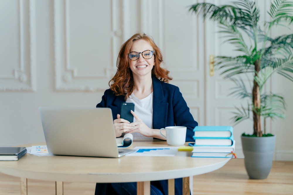 Horizontal shot of optimistic woman installs application on modern cell phone, checks notification, surrounded with paper documents, works on laptop computer, has red hair, dressed formally.