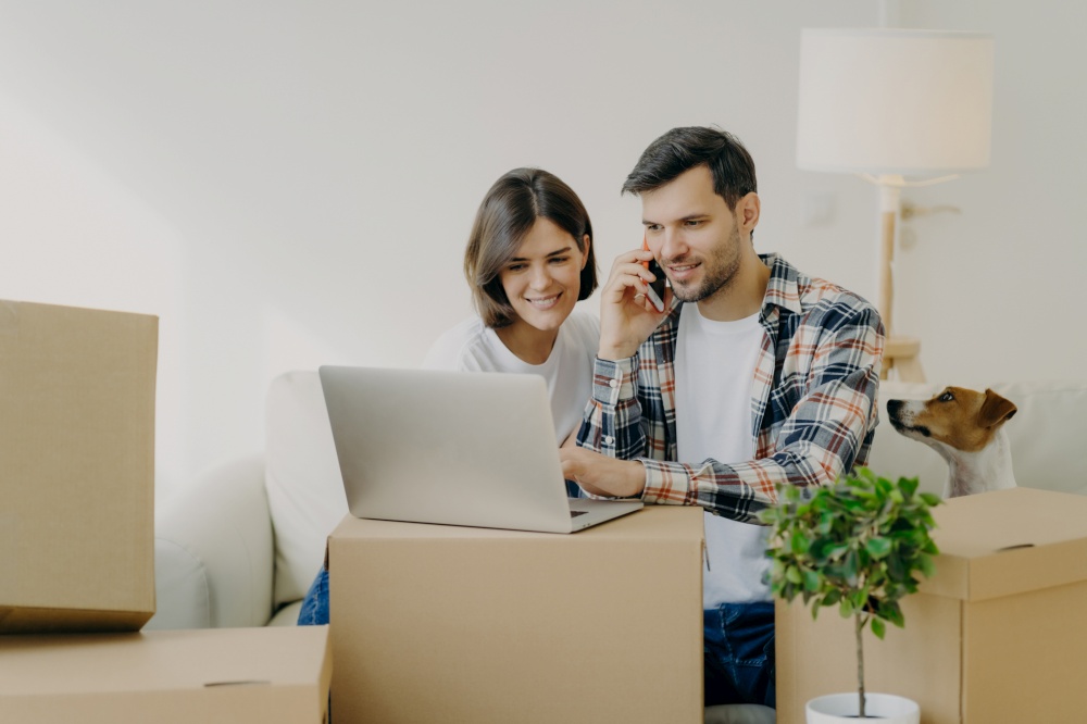 Happy man browses laptop in new flat, calls via smartphone, move in new apartment together with wife, sit on comfortable sofa, surrounded with cardboard boxes in living room, small dog near.