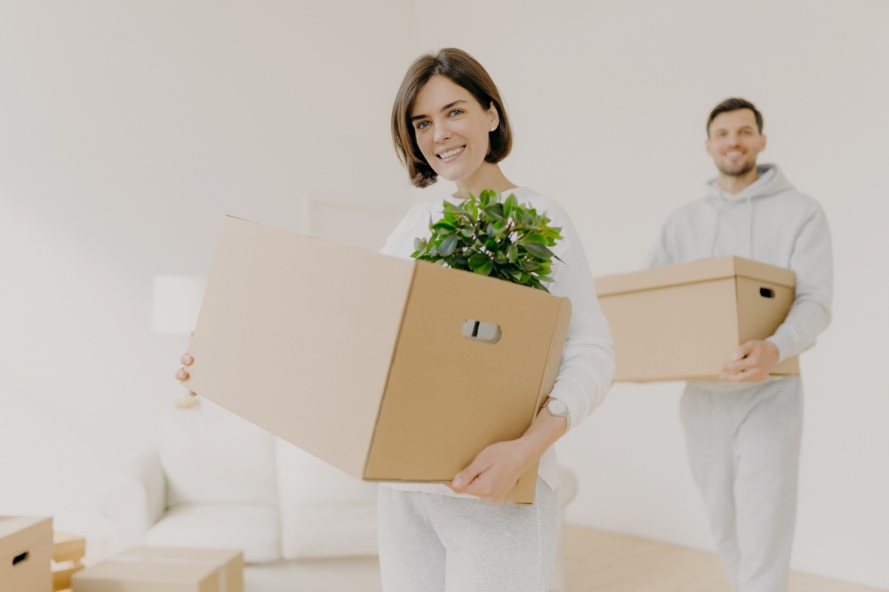 Happy young couple enter own modern house, buy real estate, carry cardboard boxes with indoor plant and other personal stuff, buy new house for living, have moving day. People and property concept