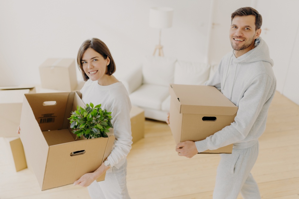 Positive female and male property owners pose with personal belongings in carton boxes, relocate to own apartment, smile pleasantly, pose in white living room. Moving day, planning, relocation