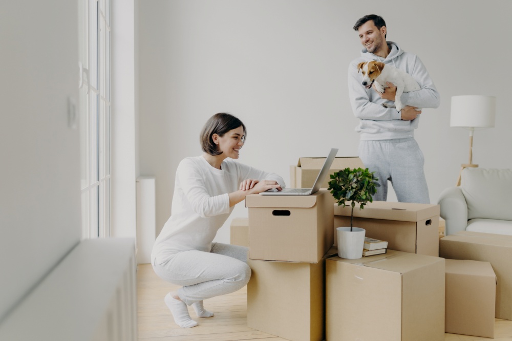 Photo of happy woman works remotely on laptop computer, has happy smile, glad husband in casual clothes plays with pet, spend free time at their new home, surrounded with cardboard boxes in middle
