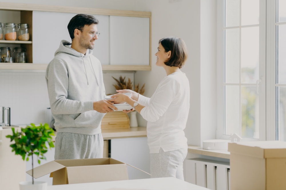 Image of delighted family couple unpack utensils with carton boxes, move in new home, pose against kitchen interior, looks happily at each other, busy unpacking different home stuff. Householding