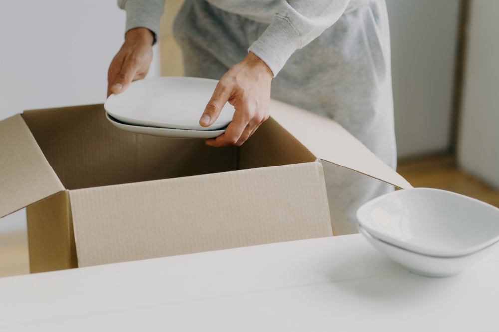 Cropped image of unknown faceless busy man unpacks boxes with dishes, holds white plates, moves in new apartment, tidies up at kitchen, has moving day. Bring out new plates. Relocation concept