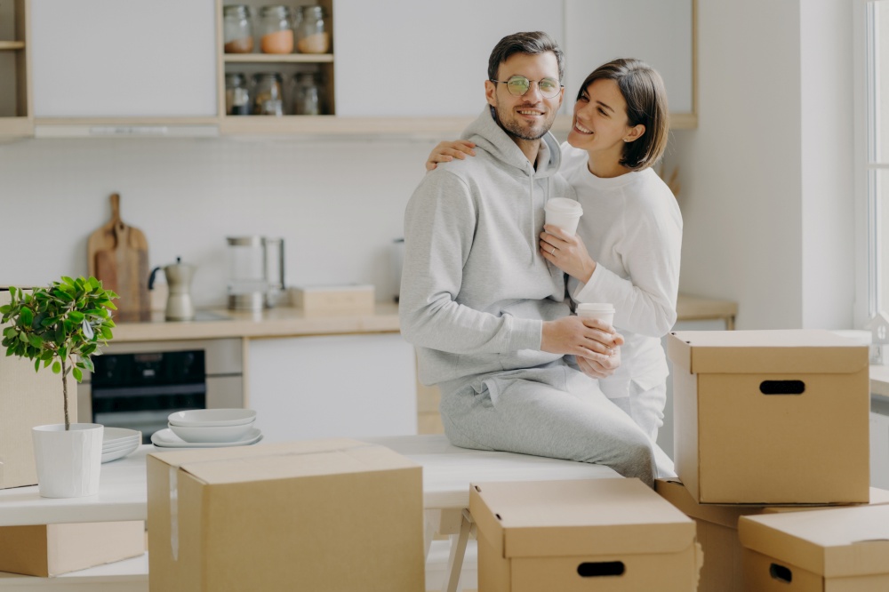 Caring woman embraces husband with love, drink takeaway coffee, pose in modern kitchen with unpacked boxes around, move into new apartment for living, rent flat, unpack belongings, have break