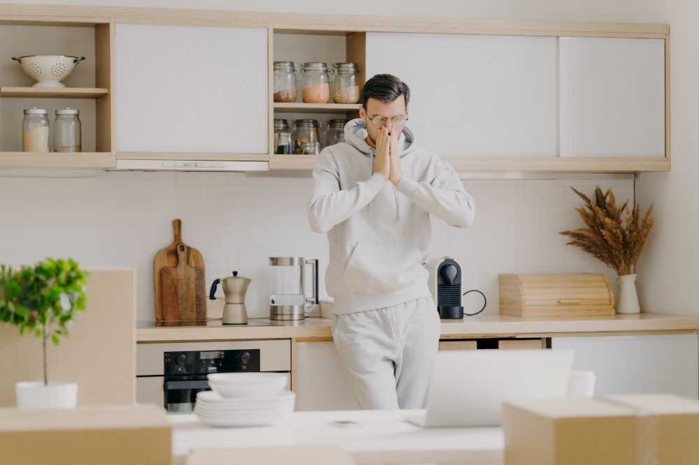 Indoor shot of pensive man works on family budget, looks attentively at laptop computer, poses in kitchen with unpacked boxes, rents new house, dressed in casual sweatshirt, works remotely at home