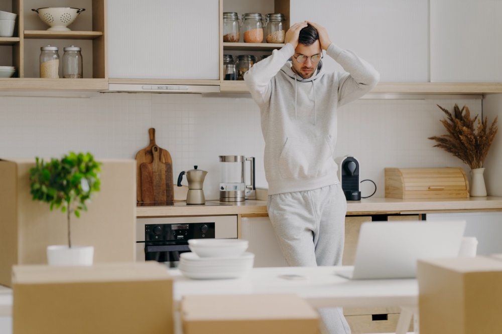 Frustrated Caucasian man faces financial problems, rents new apartment, looks with puzzlement at laptop computer, dressed in casual wear, keeps hands on head, poses against kitchen interior.