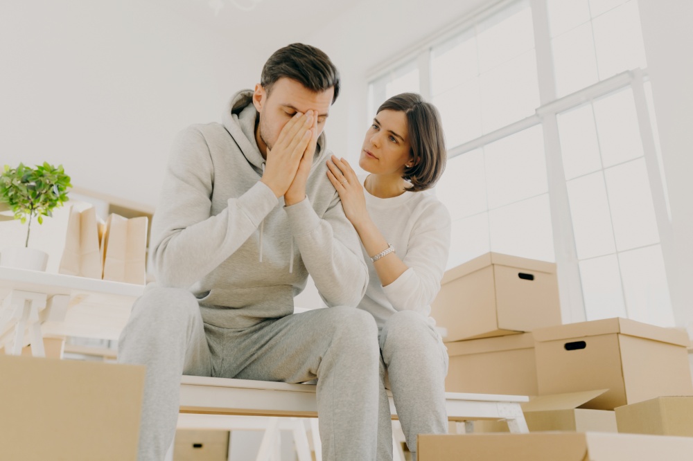 Photo of dissatisfied husband and wife forced to sell apartment because of financial problems, woman soothes husband, pose on cardboard boxes, pose in living room. Moving as kind of stress for family