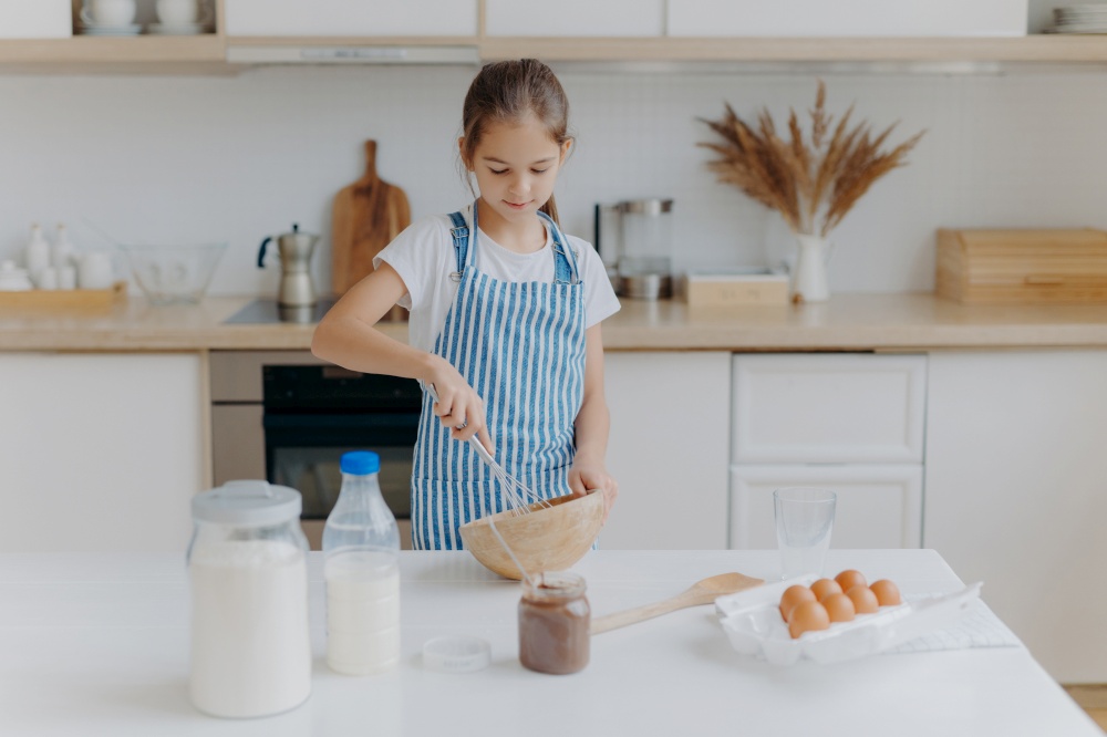 Small cute girl in apron, mixes ingredients, whisks with beater, uses eggs, milk, flour, tries new recipe, stands against kitchen interior, prepares tasty cookies or bakery, learns how to cook.