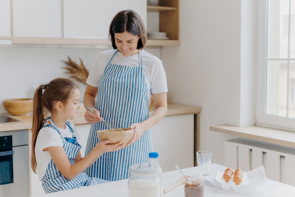 Happy family at kitchen. Lovely woman and her daughter prepare bakery together, wear aprons, like cooking together, enjoy domestic atmosphere, have fun indoor. Children, motherhood, baking concept