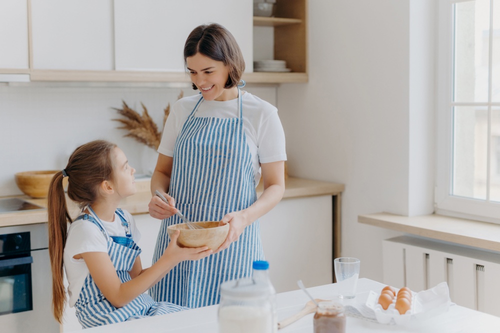 Indoor shot of caring mother with happy smile, cooks together with daughter in kitchen, prepare holiday pie, have pleasant talk, stand near table with different ingredients. Small helper helps mommy
