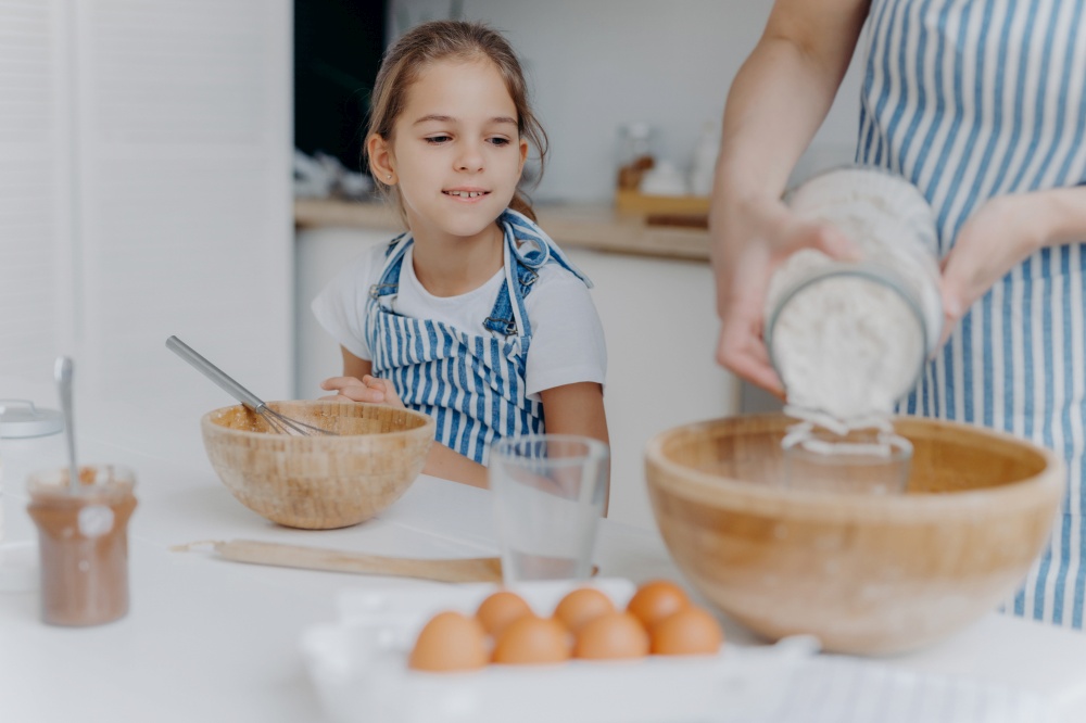 Curious little girl looks how mom prepares dough for pastry, learns to cook, gets culinary experience, wears apron. Faceless woman adds flour in bowl with other ingredients, pose in kitchen with child